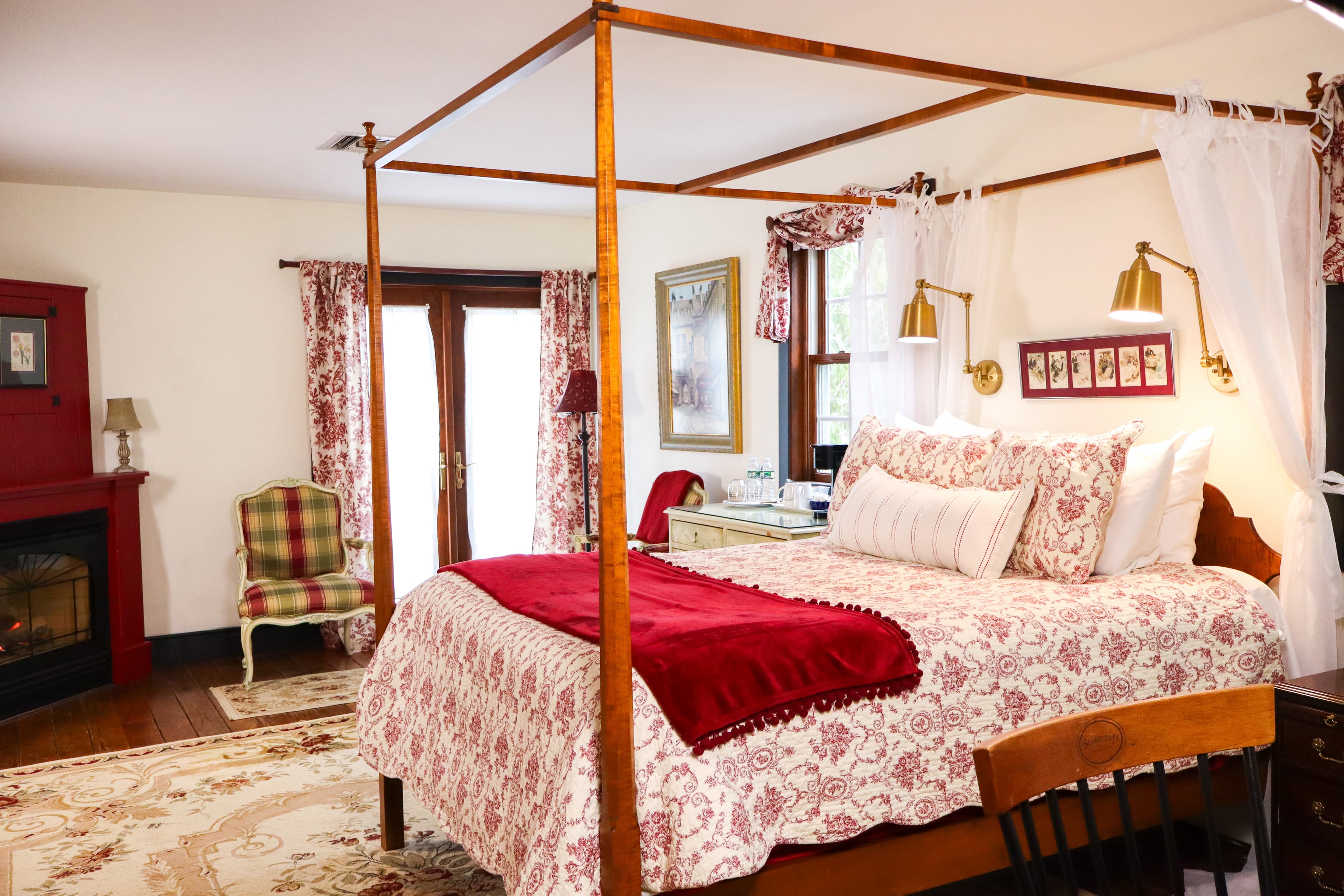 A luxurious bedroom featuring a wooden four-poster bed with sheer white drapes, a cozy fireplace, and a welcoming wine setup.