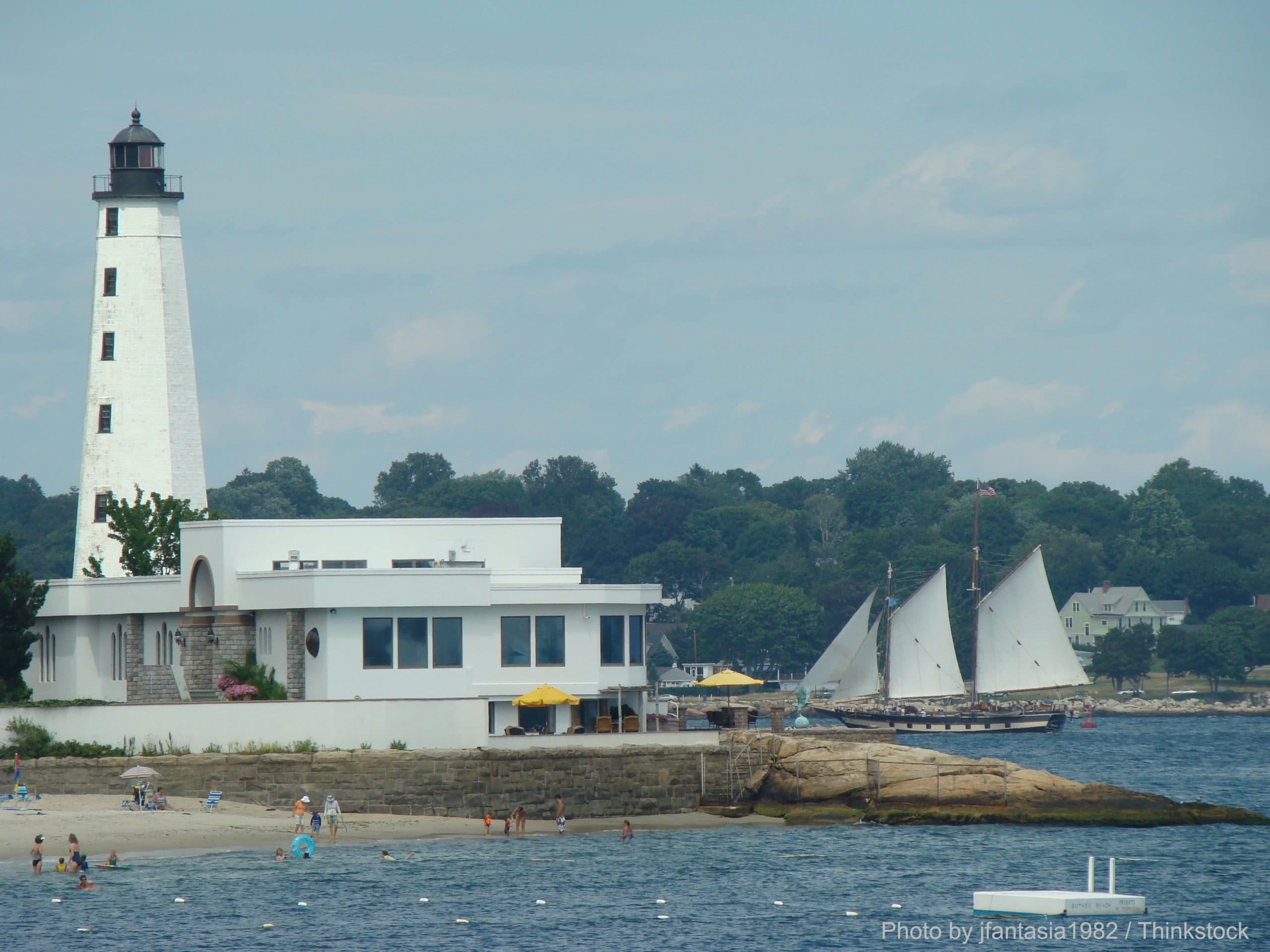 A lighthouse stands beside a sandy beach, with sailboats in the background and people enjoying the water.