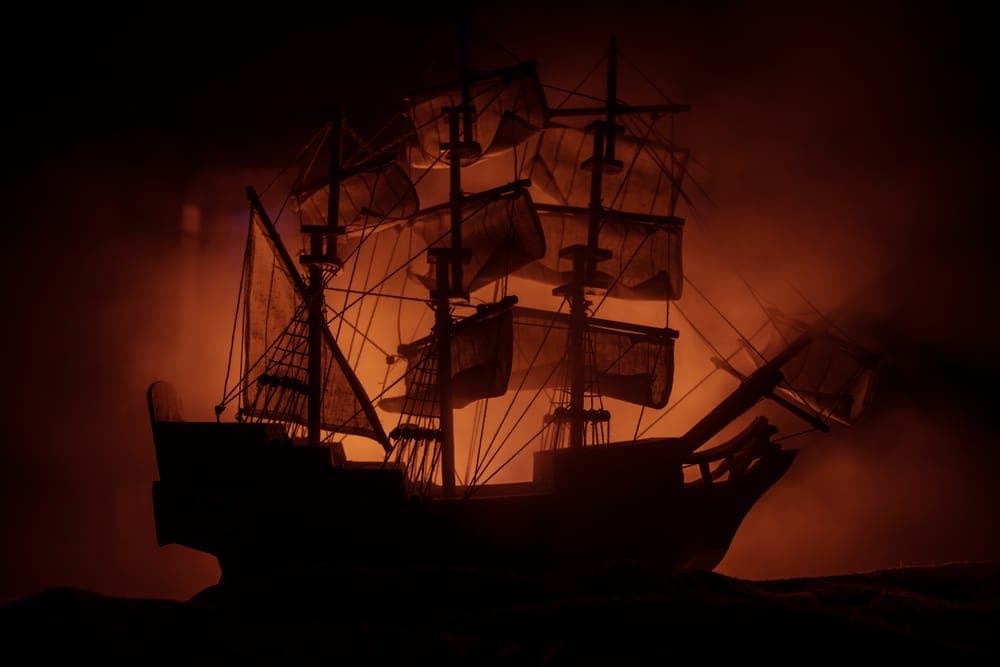 A dark silhouette of a ship with multiple sails against an orange and smoky background.