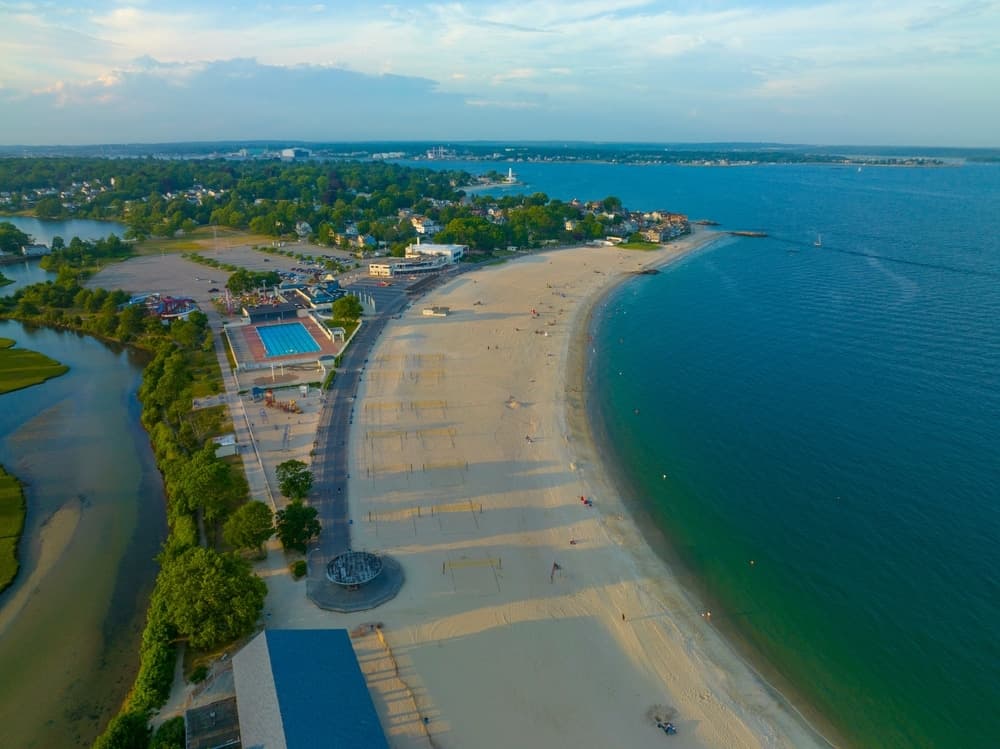 Aerial view of a sandy beach with calm waters, greenery, and towns in the background.