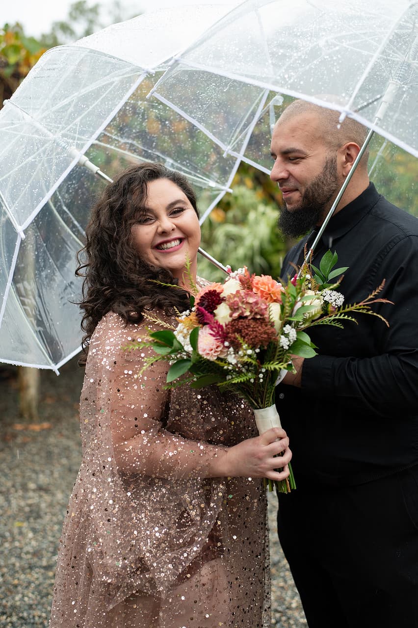 A smiling couple stands under a clear umbrella, holding a bouquet of flowers, while it rains around them.