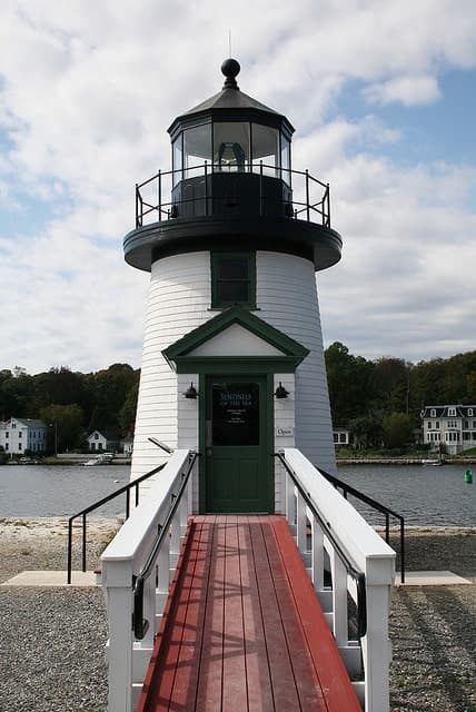A white lighthouse with a green door and red walkway beside a calm body of water.