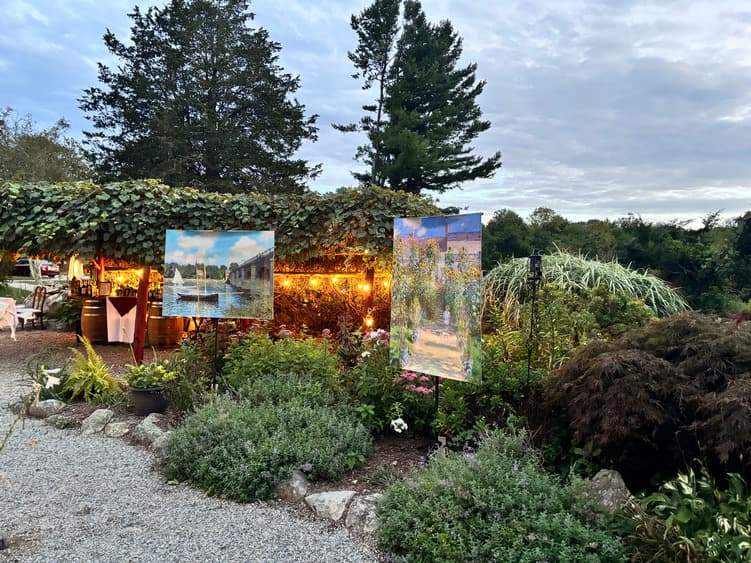An outdoor garden scene featuring two paintings on display under a vine-covered structure, surrounded by lush greenery.