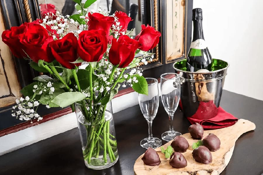 A vase of red roses, champagne flutes, and chocolate-covered strawberries on a wooden platter.