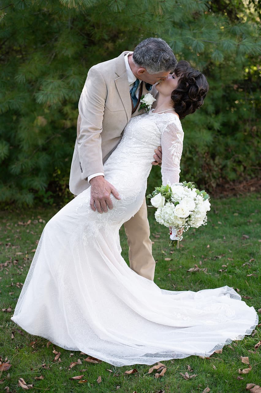 A couple shares a romantic kiss outdoors, with the bride in a white lace gown holding a bouquet of white flowers.
