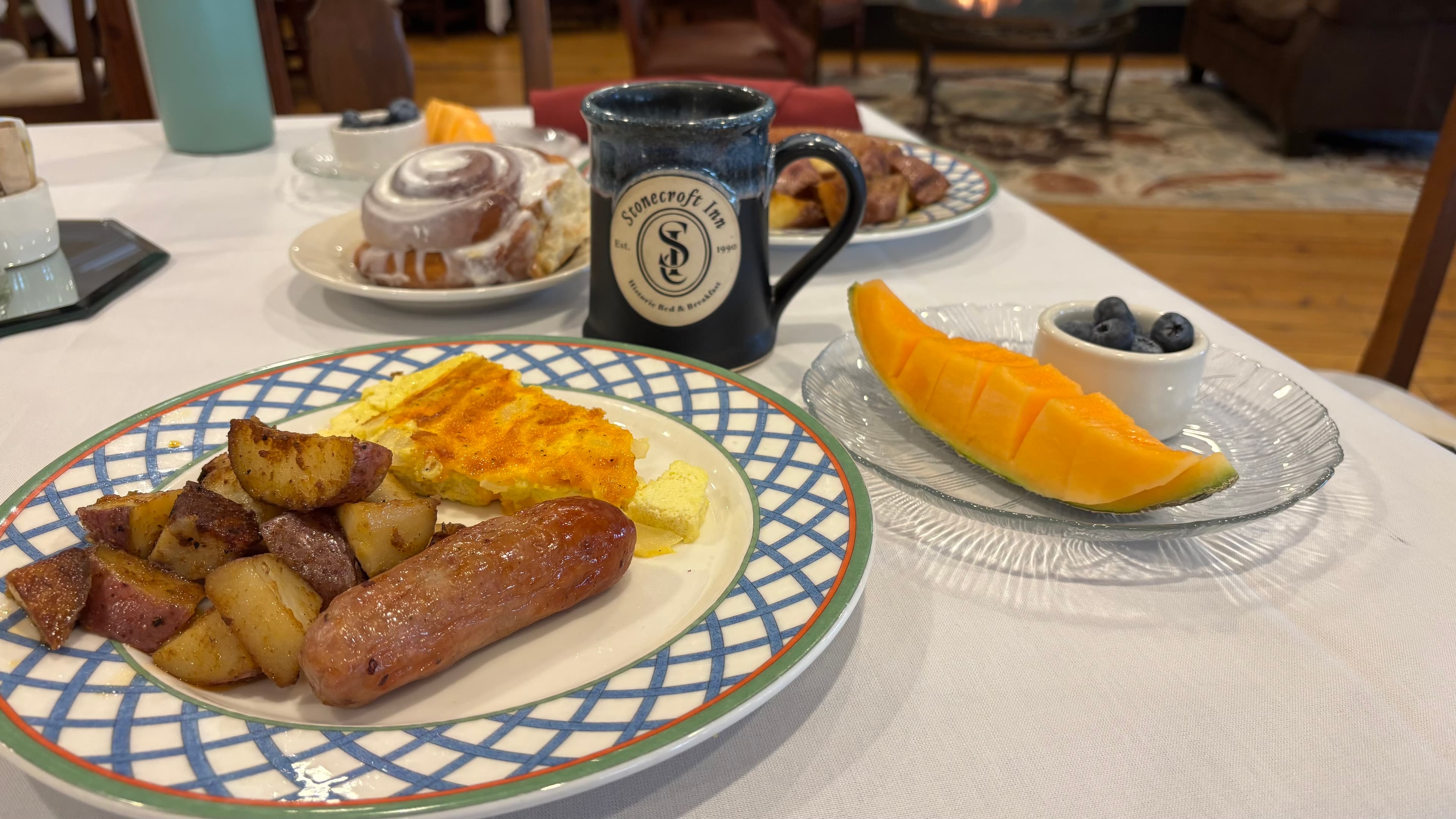 A plate of breakfast with sausage, potatoes, and eggs, accompanied by a coffee mug and a side of cantaloupe and blueberries.