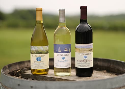 Three bottles of Stonington wine-Chardonnay, Seaport White, and Cabernet Franc-are displayed on a wooden barrel against a blurred green background.