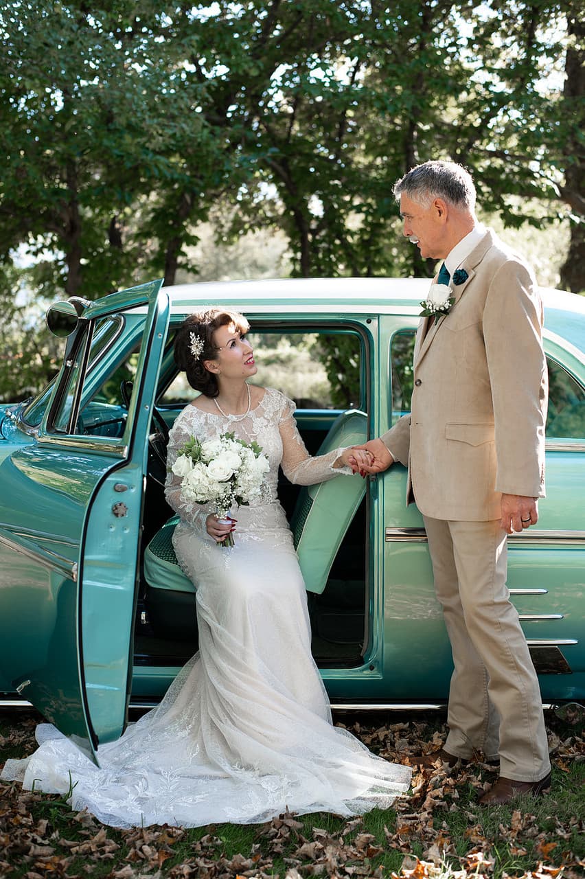 A bride in a vintage wedding dress holds a bouquet while being assisted by a man in a beige suit near a classic turquoise car.