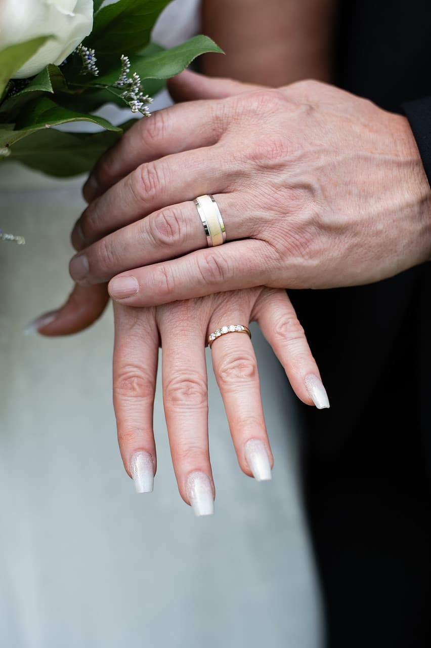 A close-up of two hands, one wearing a silver wedding band and the other adorned with a diamond ring, gently resting together.