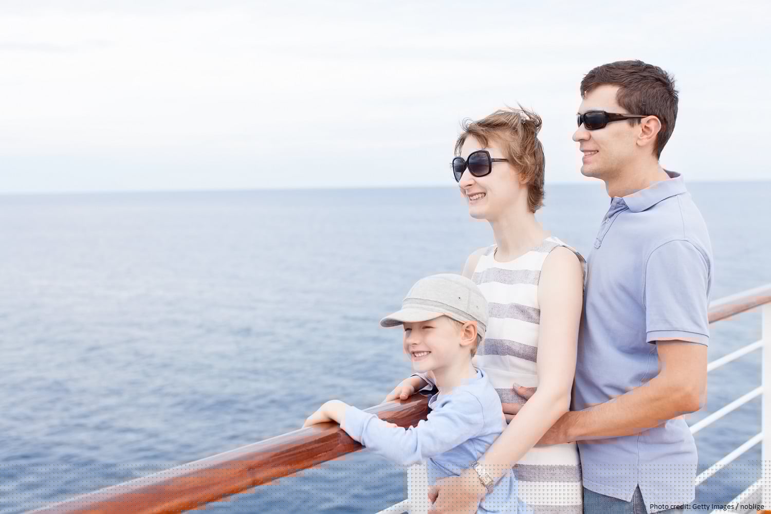 A family smiles together while leaning against the railing of a cruise ship, with the ocean in the background.