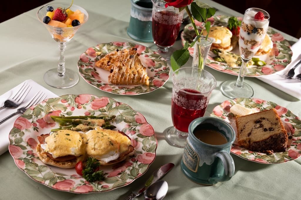 A beautifully arranged brunch spread featuring eggs Benedict, pastries, fruit, and beverages on floral-patterned plates.