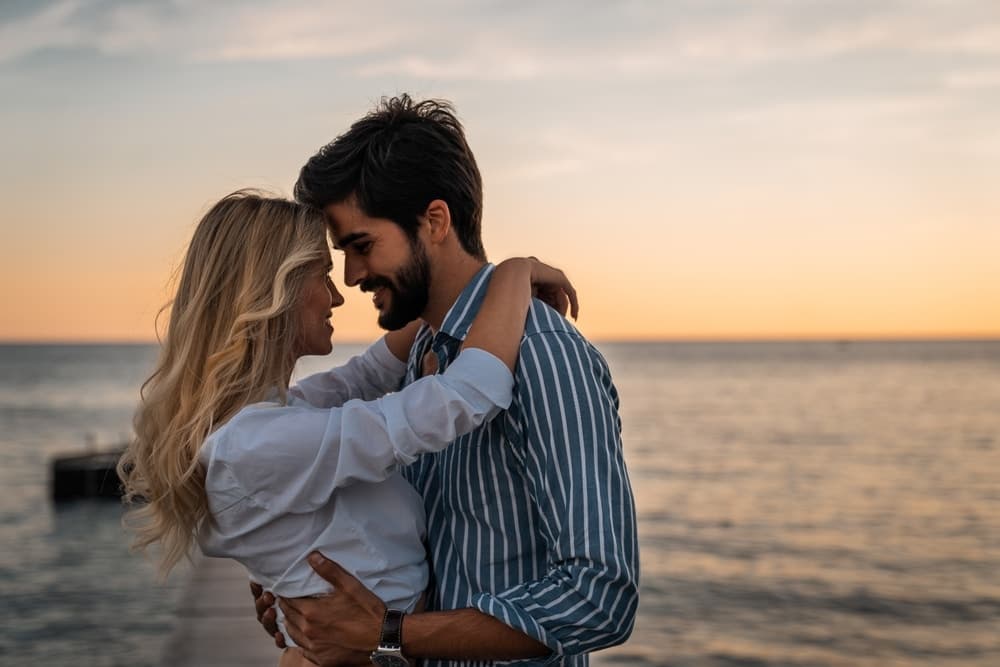 A couple embraces on a shoreline at sunset.