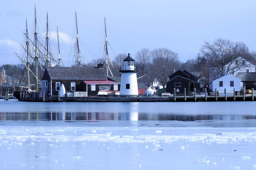 A tranquil harbor scene featuring a lighthouse and historic sailboats against a winter backdrop.
