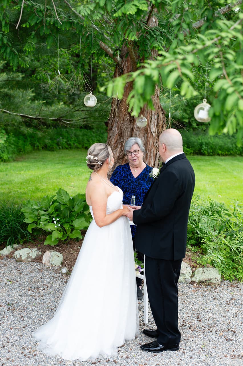 A bride and groom stand together with an officiant beneath a tree decorated with hanging ornaments.